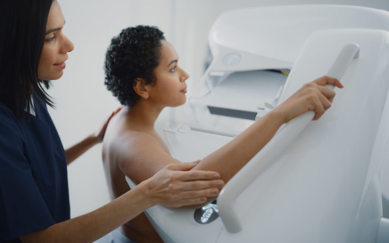 Woman holding mammogram gantry machine while mammography technologists helps her.
